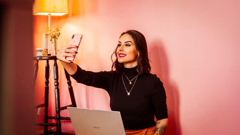 woman sitting in front of laptop and taking a video of herself using smartphone